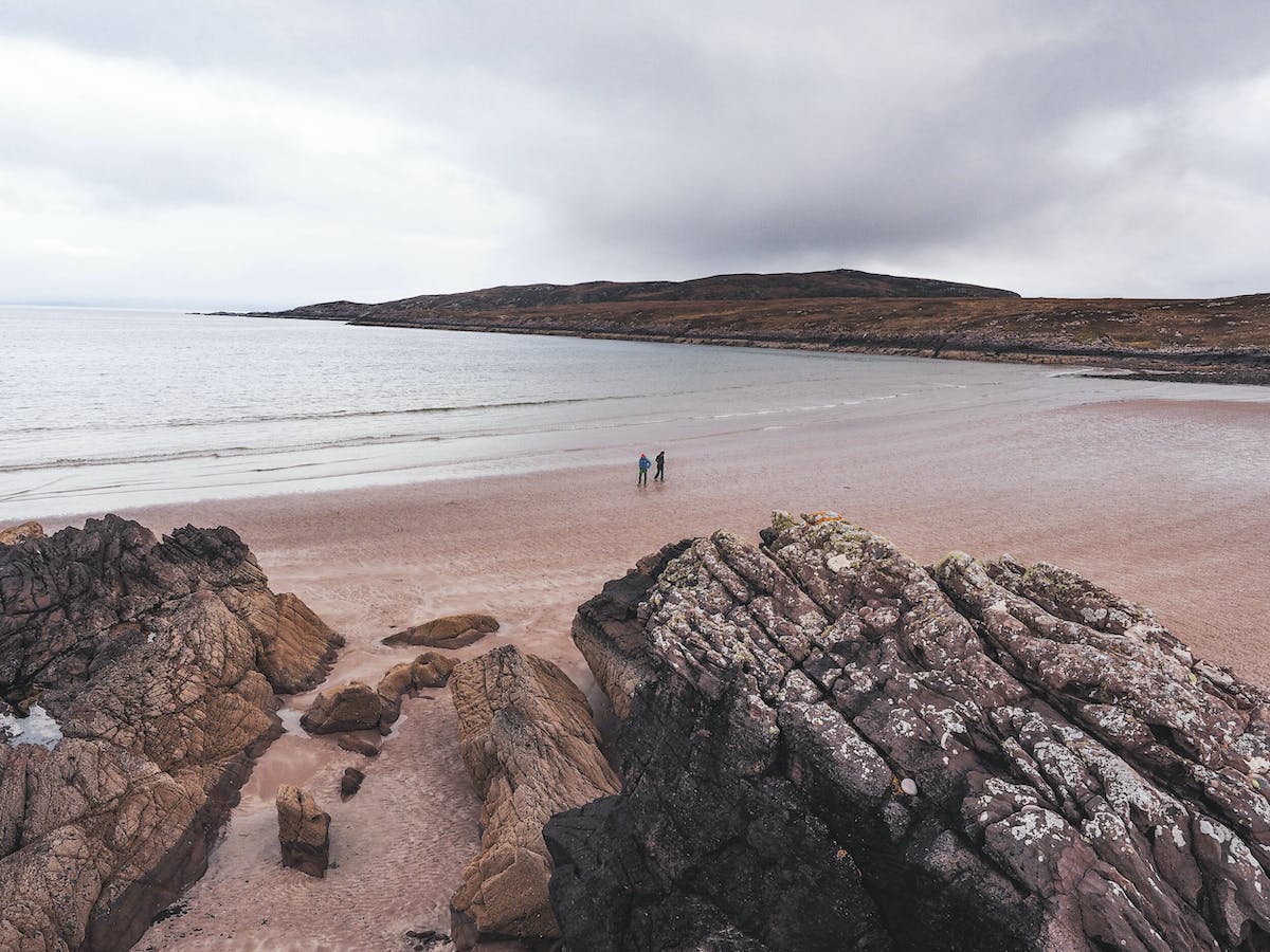 two people standing on the beach near some rocks